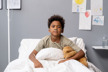 Obraz premium Portrait of African little boy looking at camera while sitting in bed with teddy bear during his treatment at hospital