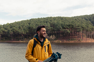 Fototapeta premium A boy with yellow raincoat standing in front of the lake. He has camp chair on his hand. He will have a rest by the lake. Nature park on a gold autumn day. He admires autumn nature landscape.