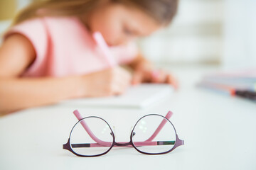 Little girl is writes in school notebook, blurry image. Glasses for vision in focus. Poor vision concept