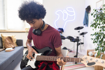 Young serious man with headphones around his neck playing electric guitar while sitting in home environment