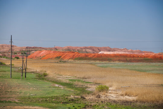 Scenery industrial landscape. Aluminium ore mining. Giant heap of red minerals, bauxite clay. Green and yellow grass on foreground. Electric poles and wires left.