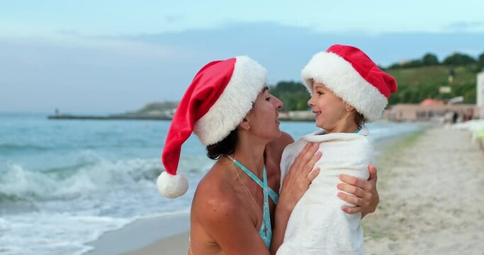 Mom and daughter are resting on the seashore during the winter holidays, a woman and a child are smiling, wearing Santa hats on their heads.