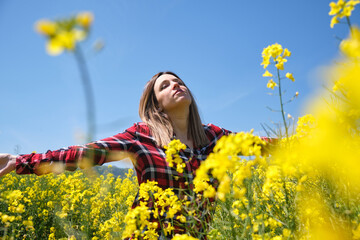 Happy woman enjoying nature, sunlight touching her face and breathing fresh air, in the middle of a beautiful field full of yellow flowers. rapeseed in spring. (Brassica napus)