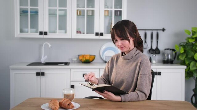 lifestyle, young woman with book in her hands reads and drinks coffee while having snack while sitting at home at table background of kitchen