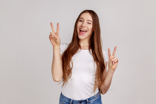 Portrait of happy attractive excited woman winking and gesturing victory sign at camera, success or achievement, wearing white T-shirt. Indoor studio shot isolated on gray background.