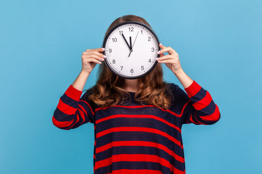 Portrait of unknown anonymous woman wearing striped casual style sweater, hiding her face, time management, schedule and meeting appointment. Indoor studio shot isolated on blue background.