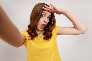 Fototapeta premium Astonished teenager girl wearing yellow casual T-shirt taking selfie, looking at camera POV, point of view of photo, looking far over head. Indoor studio shot isolated on gray background.