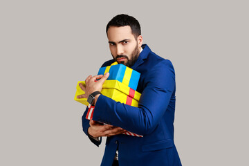 Portrait of angry serious bearded man holding stack of presents in hands, being greedy to share gifts, wearing official style suit. Indoor studio shot isolated on gray background.