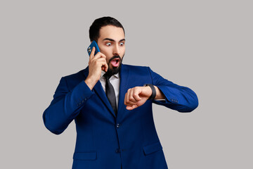 Portrait of handsome bearded man talking phone and looking at wristwatch, being late for date or business meeting, wearing official style suit. Indoor studio shot isolated on gray background.