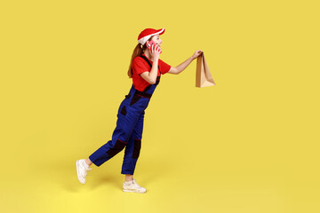 Side view portrait of delivery woman bringing door-to-door paper parcel to client and talking on phone with client, wearing overalls and red cap. Indoor studio shot isolated on yellow background.