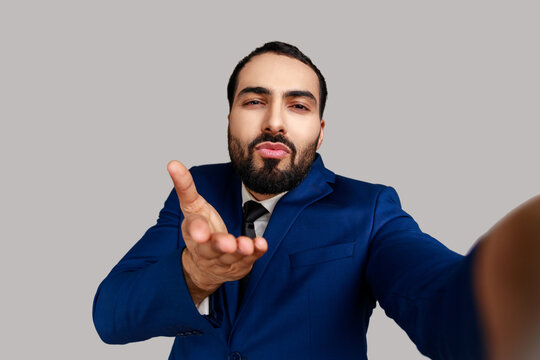 Attractive bearded man sending air kisses while taking selfie POV, flirting, expressing romantic feelings, wearing official style suit. Indoor studio shot isolated on gray background.