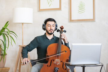 Young confident teacher of music in headphones playing string musical instrument during online...