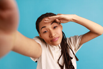 Beautiful woman with black dreadlocks has positive face expression, making selfie, looking far away, point of view photo, wearing white shirt. Indoor studio shot isolated on blue background.