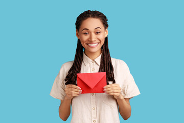Obraz premium I got love letter on Valentine's day. Happy beautiful woman with black dreadlocks holding letter in red envelope or greeting card and smiling joyfully. Indoor studio shot isolated on blue background.
