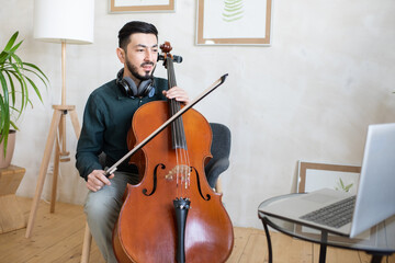 Young confident teacher with cello explaining online audience how to play it while sitting in front of laptop at home © pressmaster