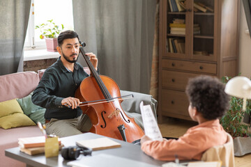 Young serious manle music teacher looking at schoolboy in front of him while playing cello during home lesson © pressmaster