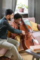 Young music teacher helping cute schoolboy playing cello while both sitting on couch in home...