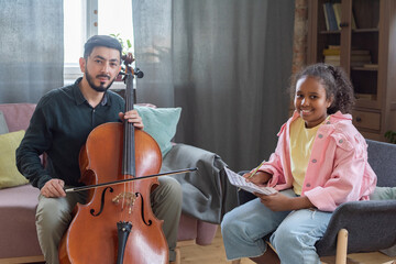 Happy little girl and her music teacher sitting in front of camera in home environment and looking...