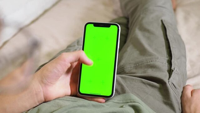 a phone with a green screen in the hands of a man lying on the bed. Modern technologies in the life of adolescents. Phone chroma. Green screen chrom key. watch from behind