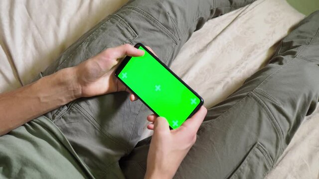 a phone with a green screen in the hands of a man lying on the bed. Modern technologies in the life of adolescents. Phone chroma. Green screen chrom key. watch from behind