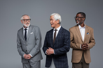 elderly multiethnic businessmen in formal wear smiling at each other isolated on grey