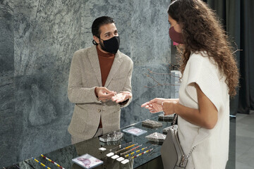 Young man in protective mask helping female client to choose bracelet or necklace while both...