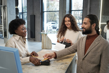 Happy young female shop assistant serving male client paying by card for the goods purchased in...