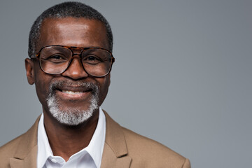 bearded african american businessman in eyeglasses smiling at camera isolated on grey