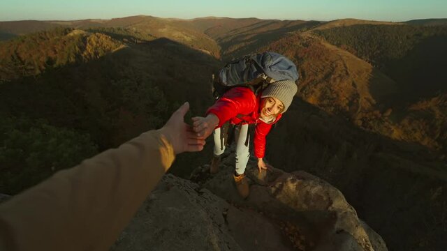 Close up of male's hand stretching out and helping female hiker reach cliff top by pulling her up from edge. POV of man giving hand to woman climbing on cliff and helping her reach top. autumn hike.