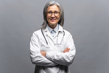 Smiling gray-haired female doctor pediatric, physical, therapist wearing white medical gown with stethoscope on shoulders stands with arms crossed isolated on gray, copy space. Healthcare and medicine