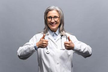 Cheerful senior mature female doctor with a stethoscope in a medical gown posing isolated over gray background, showing a thumbs-up. Smiling and welcoming healthcare worker looking at the camera