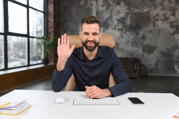 Smiling bearded caucasian businessman sitting at the desk and looking at the camera while waving....