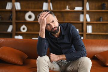 Full of doubts caucasian guy sitting on the sofa at home. Frustrated pensive man worried about future, covering forehead with hand, thinking about troubles and problems