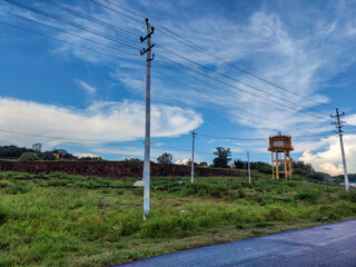 Obraz premium Stock photo of empty roadway along with electric pole with power transmission line on the left side of the road. old rusty water tank tower situated in the corner of the grassland at Gulbarga, India.