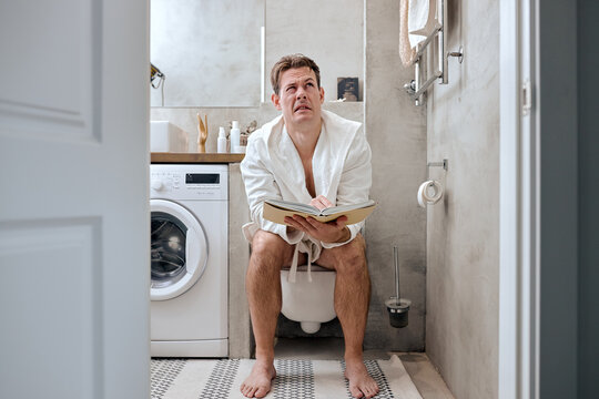 Male sitting in toilet feeling unhappy and holds book, diarrhea constipation,Health concept. young adult caucasian male in toilet in the morning at home. Hygiene at Morning Concept.