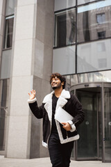 Positive indian man in jacket holding laptop and waving on urban street 