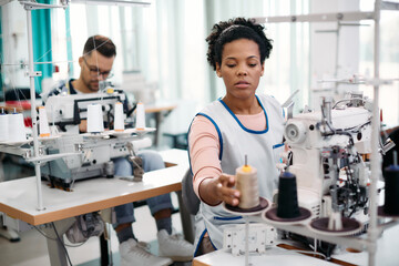 Black seamstress adjusts thread on sewing machine while working at textile factory.