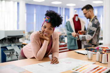 Happy black clothing designer works at her fashion design studio and looks at camera.