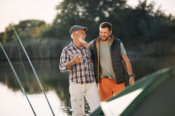 Cheerful father and son talk while fishing and camping by lake.