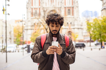 Curly indian tourist with backpack using cellphone on urban street 