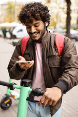 Young indian man using smartphone while renting electric scooter outdoors 