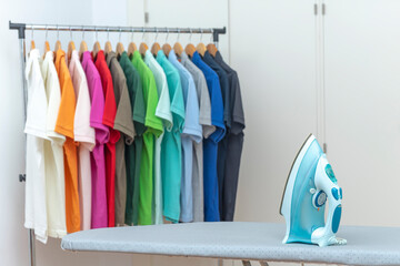 Iron on the ironing board and many colored polo shirts hanging on the rack in the background. Housework concept.
