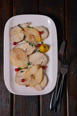 Thin slices of baked chicken breast, slices of pear, viburnum berries on a white rectangular dish, wooden background.