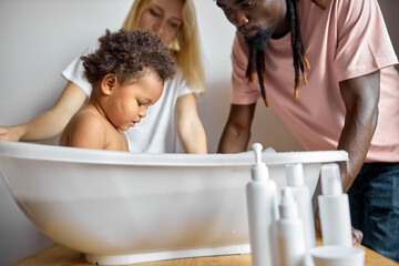 charming black child girl with curly hair sitting in bathtub, parents washing daughter in foam....