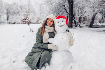 Woman hugs snowman in snowy winter park dressed in Christmas Santa hat. Festive fun outdoor...