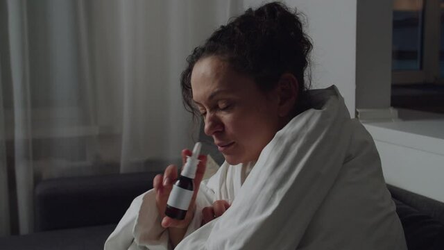 Portrait of sick adult woman sitting on couch wrapped in blanket around her, using nasal spray, applying remedy into each nostril indoors. Attractive lady has flu, cold or virus, treating at home