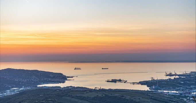 Aerial or elevated view of colorful sunset above Adriatic Sea and Trieste city. Time lapse of colorful clouds moving during golden hour. Coastline in the foreground. Romantic summer evening
