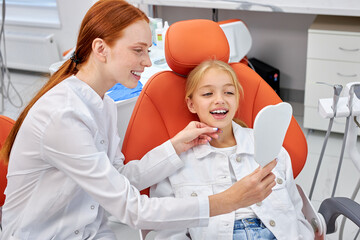 Redhead professional woman dentist in uniform works with child patient in dental office, showing patient's teeth on mirror, confidently explaining how to treat and which tooth will be after treatment