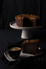 A big chocolate cake slice and a cup of coffee over a ceramic tray with a blurred cake as background.