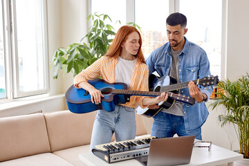 Handsome man in casual wear teaching girlfriend to play the guitar. Guy shows woman how to play guitar. Couple in casual wear using musical instrument, laptop and piano. In bright cozy room